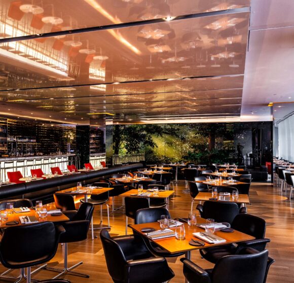 Modern restaurant interior with sleek, reflective ceiling, wooden floors, and black leather chairs around wooden tables. A long bar area with high chairs is visible to the left. Soft, warm lighting illuminates the space, creating an inviting atmosphere. - Hausion