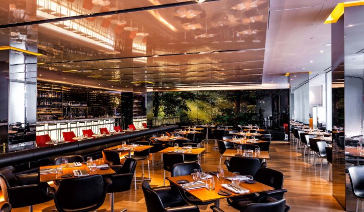 Modern restaurant interior with sleek, reflective ceiling, wooden floors, and black leather chairs around wooden tables. A long bar area with high chairs is visible to the left. Soft, warm lighting illuminates the space, creating an inviting atmosphere. - Hausion