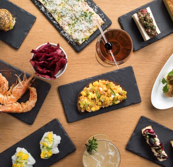 A variety of gourmet dishes arranged on a wooden table. Plates of shrimp, mini burgers, toast with toppings, meatballs, a flatbread, and cocktails are artfully displayed on black slate serving trays, showcasing diverse flavors and vibrant colors. - Hausion