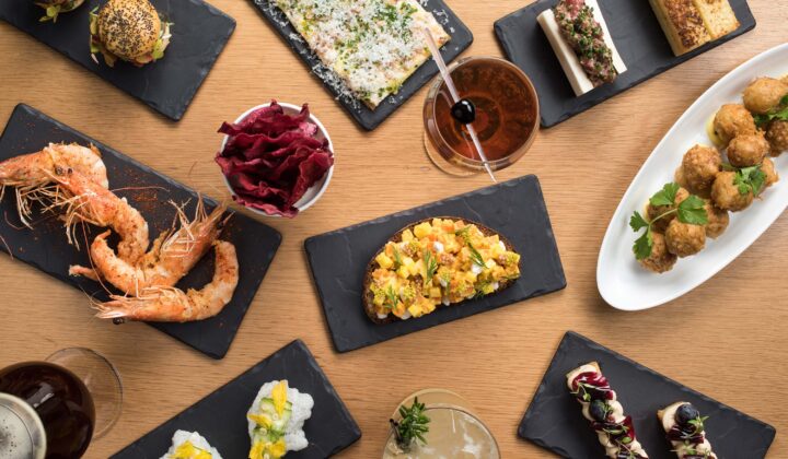 A variety of gourmet dishes arranged on a wooden table. Plates of shrimp, mini burgers, toast with toppings, meatballs, a flatbread, and cocktails are artfully displayed on black slate serving trays, showcasing diverse flavors and vibrant colors. - Hausion