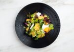 A gourmet salad served on a black plate, featuring a vibrant mix of leafy greens, thinly sliced citrus fruits, possibly oranges, and garnished with small round green elements. The dish is aesthetically presented against a marble surface. - Hausion