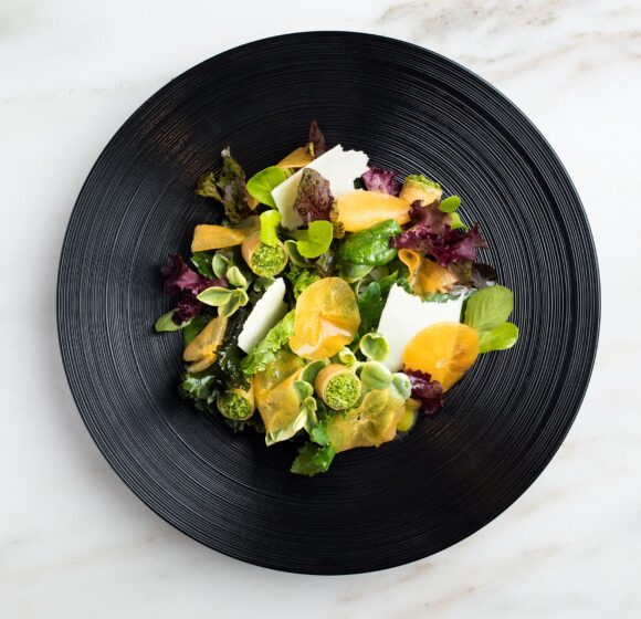 A gourmet salad served on a black plate, featuring a vibrant mix of leafy greens, thinly sliced citrus fruits, possibly oranges, and garnished with small round green elements. The dish is aesthetically presented against a marble surface. - Hausion